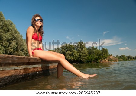 Pretty smiling teenage girl sunbathing while sitting on river boat on hot summer day.