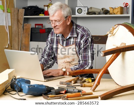 Portrait of retired carpenter sitting at his workshop and working with laptop next to the broken legged chair. Small business. 