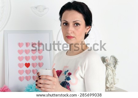 A young beautiful woman holding a white candle. In the background is a picture with hearts and angel statuette. Her hair is pulled back in hairstyle.