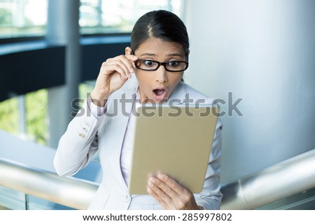Closeup portrait, funny young woman, astonished surprised, wide open mouth, large eyes in black glasses by what she sees on her gray silver tablet pad, isolated indoors office background.