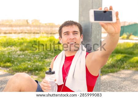 Sitting guy in park and take a selfie during a break at run training with friends - listen music with headphones - Young man finish to do sport and make himself picture - fitness jogging workout 