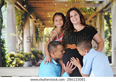 A happy  mom-to-be-again and her young daughter with the dad and elementary son touching and kissing her pregnant belly.  