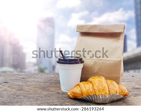 coffee and croissant with paper bag on wooden table over cityscape blur background