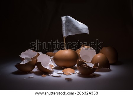 Conceptual image, Broken brown eggs with one unbroken, one white flag raise up from the egg as surrender. Funny image of eggs with spot light, black background. Lose from the beginning.