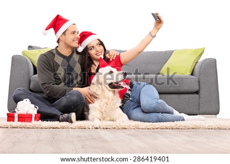 Young couple with Santa hats taking a selfie with their dog seated in front of a modern gray sofa isolated on white background