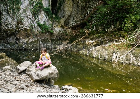 Tired and annoyed little girl resting on a big rock by a small pond, during a forest hike, exploring big nature. Active, healthy and natural lifestyle concept. 