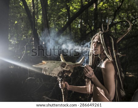 portrait of woman with long dreadlocks hair hold in hand staff with cow skull with horn against  wild forest trees Young girl smoke cigarette Woman shaman in ritual garment stand from fur and leather