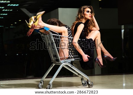 Two young misterious funny fashionable girls in dresses with shopping trolley indoor on store backdrop, horizontal picture