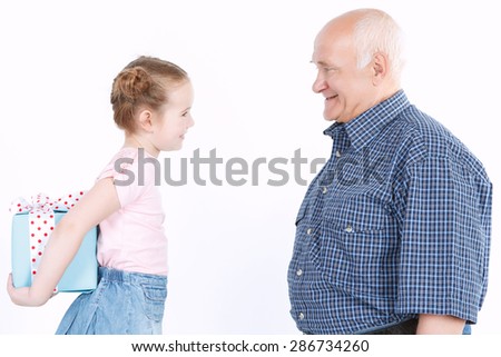 Portrait of a small pretty granddaughter standing in front of her smiling grandfather and hiding a present behind her back preparing making a surprise, isolated on a white background
