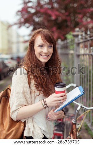 Pretty young university student standing alongside her bicycle in an urban street holding her notes and a cup of takeaway coffee with a backpack over her shoulder