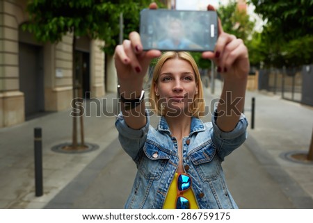 Gorgeous young woman making self portrait with a cell phone camera while enjoying a day out strolling in new city at summer vacation, stylish female hipster taking a picture of herself on smart phone