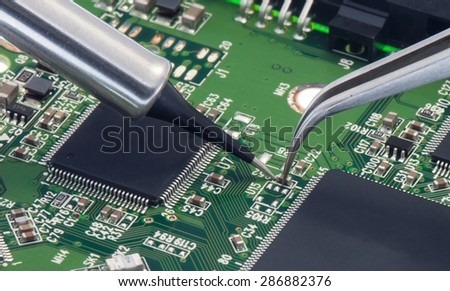 Electronic component  held with tweezers by an engineer over a green motherboard.