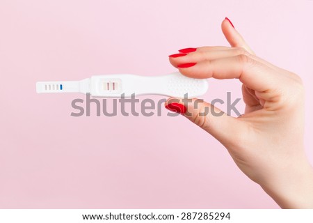 Beautiful female hand with red fingernails holding positive pregnancy test isolated on pink background. Motherhood, pregnancy, birth control concept. Minimal sparse modern image language.  Royalty-Free Stock Photo #287285294