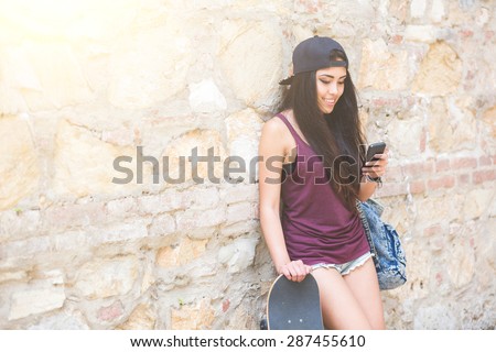 Portrait of a beautiful skater girl looking at smart phone against stone wall. She is half caucasian and half filipina, she wears short jeans, a purple tank top and a black cap.