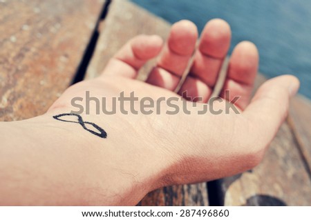 closeup of the arm of a young caucasian man with an infinity symbol tattooed in his wrist, on the boardwalk of a dock