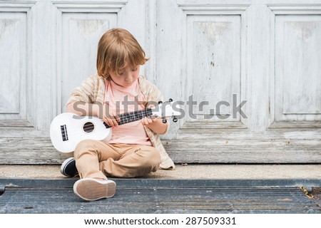 Little happy boy plays his guitar or ukulele, sitting by the wooden door outdoors Royalty-Free Stock Photo #287509331