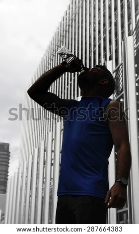 Silhouette of young sport man drinking water bottle after running training session in business district with office buildings and blue sky background in healthy lifestyle and fitness concept