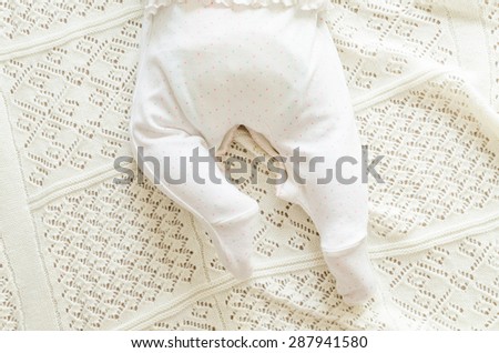 Tiny little newborn daughter baby's feet in nice white spotted romper suit lying on the background of white woolen blanket on a bed at home, top view. 