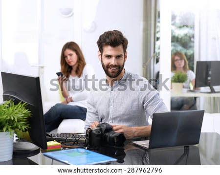 Portrait of young journalist sitting at editorial office in front of laptop and computer and working online. Assistant working at background. 