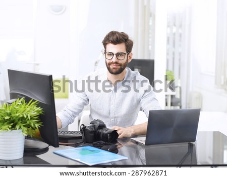 Portrait of young journalist sitting at editorial office in front of laptop and computer while smiling and looking at camera.