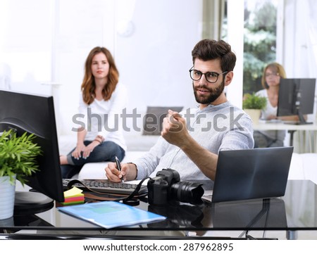 Portrait of young graphic man sitting at studio in front of laptop and computer while working online. Assistant working at background. Small business. 