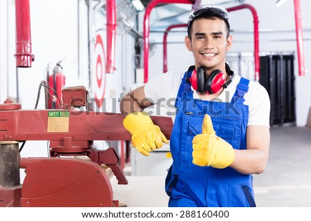 Proud and successful Asian carpenter standing next to circular saw giving thumbs up sign 