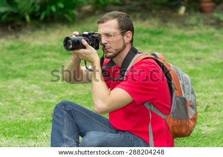 Man in red t-shirt taking a photo in the park with grassy background