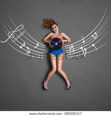 Beautiful pin-up girl in retro bikini and sunglasses, holding an LP microgroove vinyl record and sitting on musical staff chalk drawings background, top view.