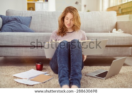Pretty young girl doing her studies at home sitting on the floor in the living room with a laptop computer reading a binder of class notes Royalty-Free Stock Photo #288630698