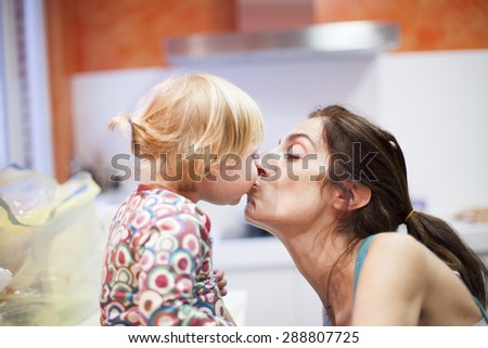 nineteen month aged blonde baby colored shirt with pigtails and brunette woman mother kissing in mouth in orange kitchen