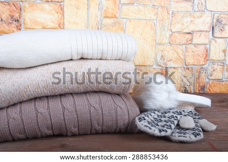 Mittens, fur headphones and stack of knitting clothes on wooden table opposite a stony wall.