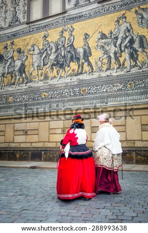 two young girls in traditional old costumes under detail of giant antique mosaic picturing monarchs over the centuries, in the streets of Dresden, Germany