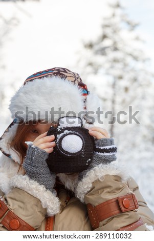Beautiful young woman in winter clothing, fingerless mittens and ornamented hat photographing snowy forest by a knitted camera. Shallow dof. Focus on hands and camera.