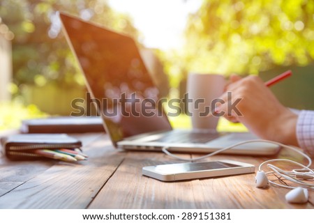 Freelance work.
Casual dressed man sitting at wooden desk inside garden working on computer pointing with color pen electronic gadgets dropped around on table side view Royalty-Free Stock Photo #289151381