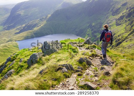 A female hiker walking down Long Stile towards Rough Crag with Blea Water in the distance in the Lake District. Royalty-Free Stock Photo #289258370