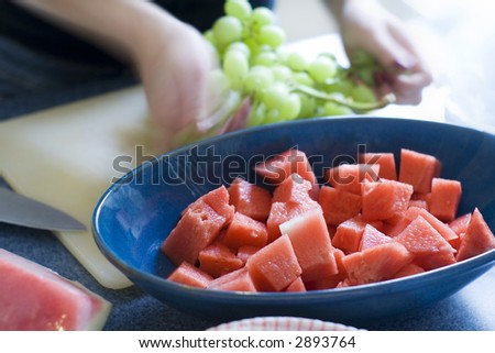 A blue bowl of chopped water melon on a counter top. A woman prepares food in the background. Slow shutter speed on tripod to capture blurred motion.