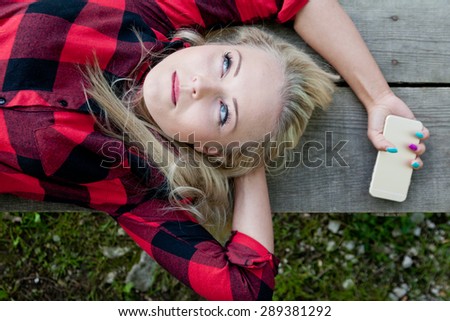 blonde beautiful girl thinking or realizing about something on the bench of a park with her mobile in her hand
