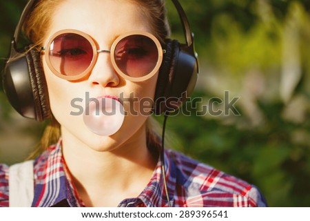 Young hipster girl listening to music on headphones in a summer park. Portrait close-up with chewing gum. Warm toning. Concept of cheerful youth. Royalty-Free Stock Photo #289396541