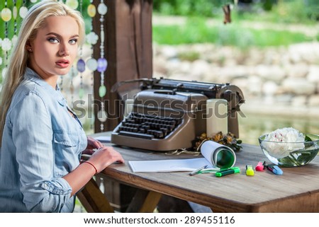 Beautiful young blonde sits in arbor at oak table near vintage typewriter & makes entries in sheets of paper. 