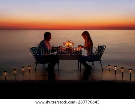 Loving couple share a romantic dinner with candles and lanterns light way at sea beach in water against wonderful sunset Royalty-Free Stock Photo #289790645