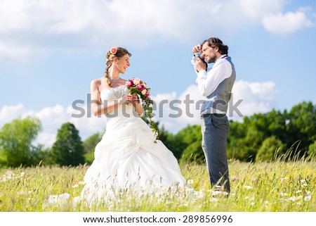 Wedding groom photographing bride with camera outside on field or meadow