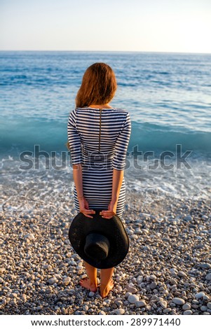 Young and elegant woman in stripped dress with a hat standing on the beach on sunset. Back view