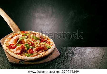 Ham, tomato and arugula, or rocket, pizza served at a pizzeria or restaurant on a long handled wooden board on a rustic counter with copyspace behind