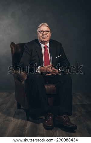 Friendly successful senior businessman wearing eyeglasses sitting forwards in an armchair in a shadowy room looking at the camera with a smile