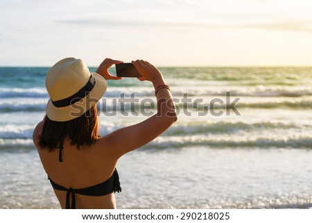 Bikini asia woman tourist taking photo on sea beach with mobile smart phone camera at sunset