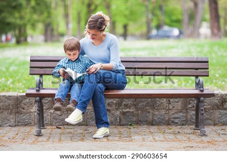 Mother and son sitting on a bench in a park and reading a book