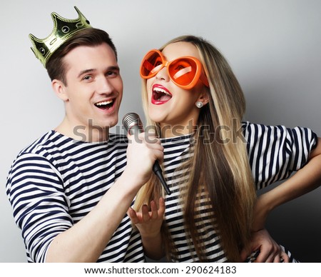 people, friendship, love and leisure concept - beautiful young loving couple with microphone. Big orange glasses and crown. Ready for party.