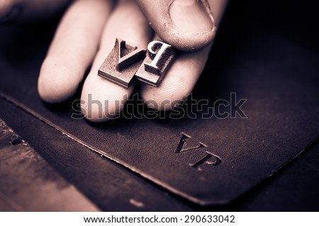 Working process of making leather wallet in the leather workshop. Vintage monogram emblem with letters V and P. Craftsman's hand holding the iron patters. Black and white photography. Cream toned.