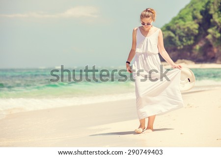 Young woman in long white dress with hat in her hand walks along tropical beach having great summer time on holidays. Summer vacation, holidays, serenity, travel, lifestyle concept 
