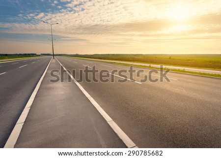 Driving on an empty asphalt six lane highway through the cultivated fields towards the setting sun. Royalty-Free Stock Photo #290785682